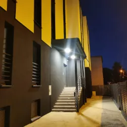 Zewnętrzne oświetlenie budynku z czarno-żółtą fasadą i schodami prowadzącymi do wejścia, widoczne z boku, oświetlone lampami zewnętrznymi w nocy.