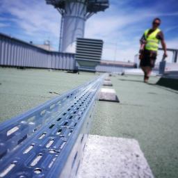 InSE-ET Jarosław Kowalczyk - Metalowa korytka kablowa ułożona na betonowych wspornikach na dachu budynku, w tle pracownik w kamizelce odblaskowej i konstrukcja wieżowa.