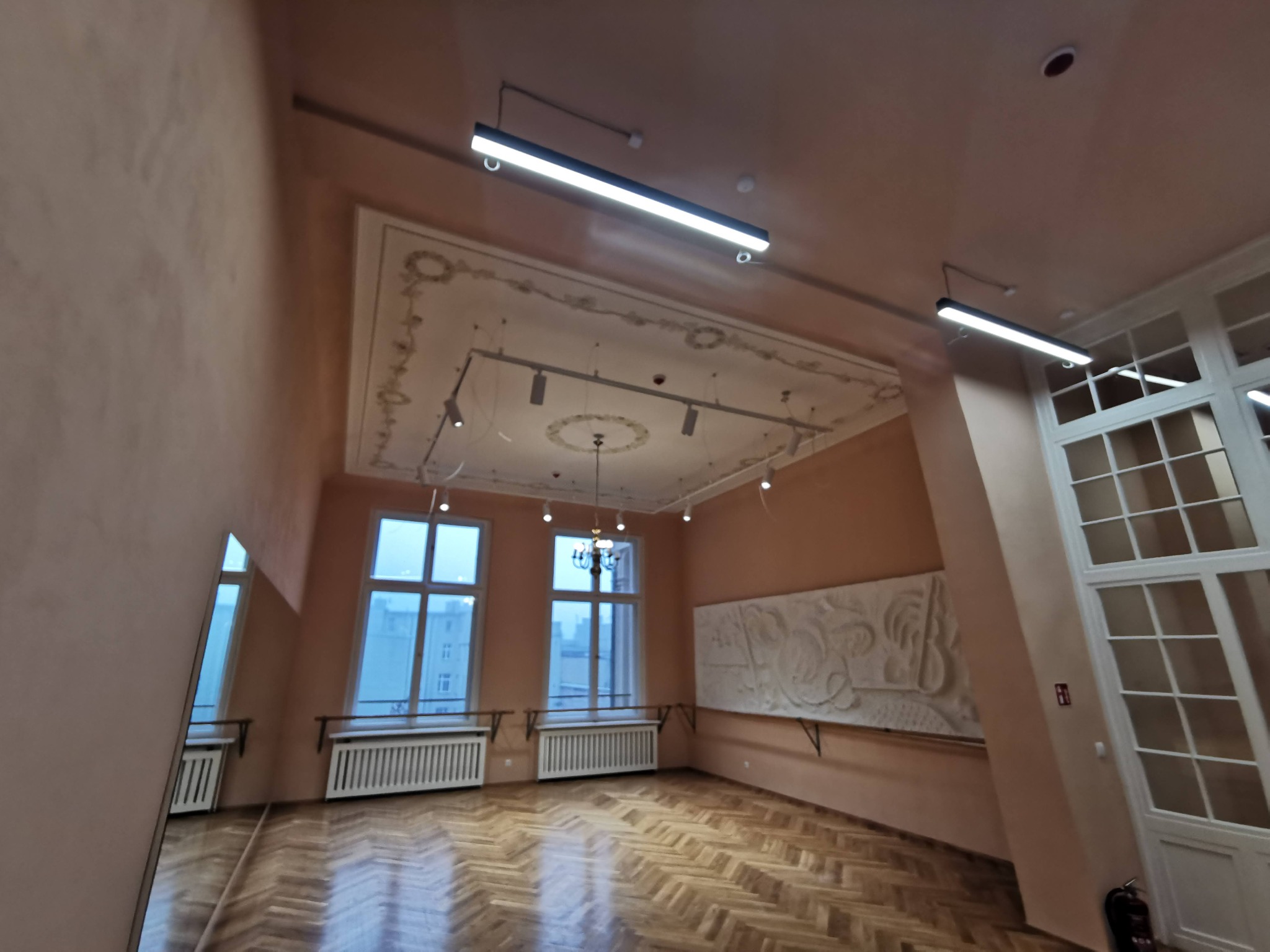 Wnętrze sali z parkietem w jodełkę, oświetleniem liniowym LED i ozdobnym plafonem, widoczne okna i płaskorzeźba na ścianie.