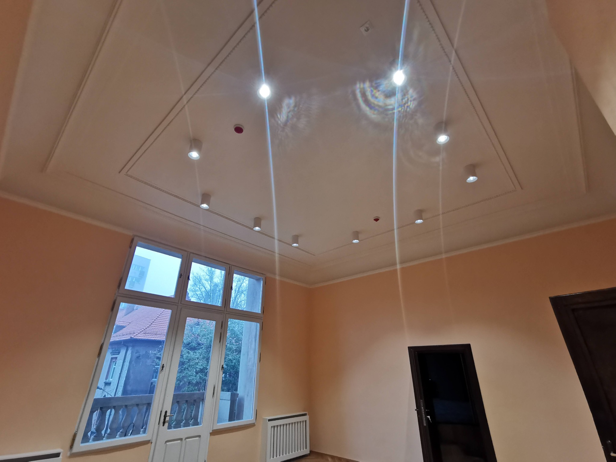 Wnętrze pokoju z zamontowanym oświetleniem punktowym w ozdobnym suficie podwieszanym, widok na okno balkonowe z widokiem na budynek mieszkalny i drzewa.