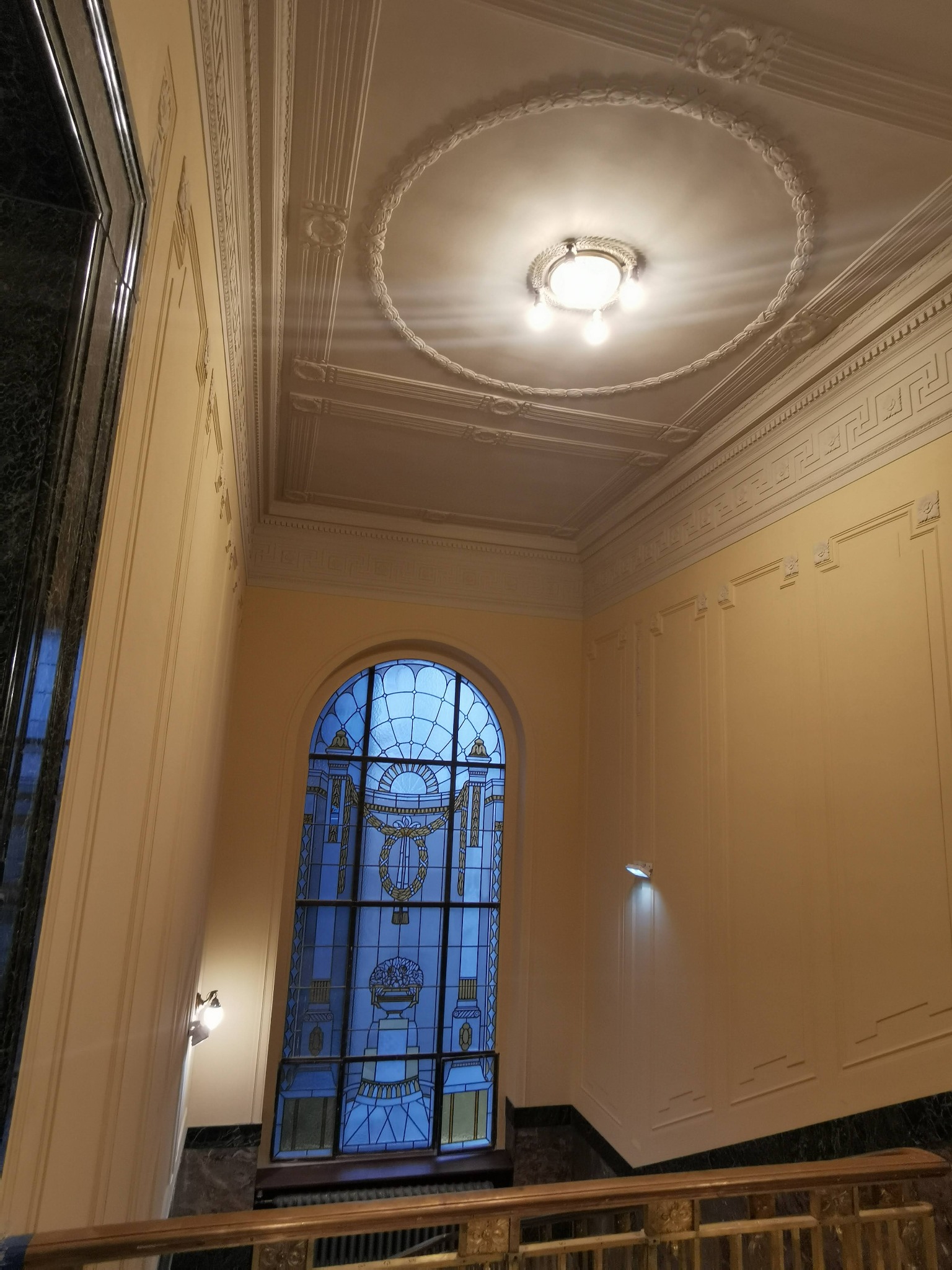 Wnętrze klatki schodowej z wysokim witrażowym oknem, ozdobnym sufitem z okrągłym żyrandolem i kinkietami ściennymi.