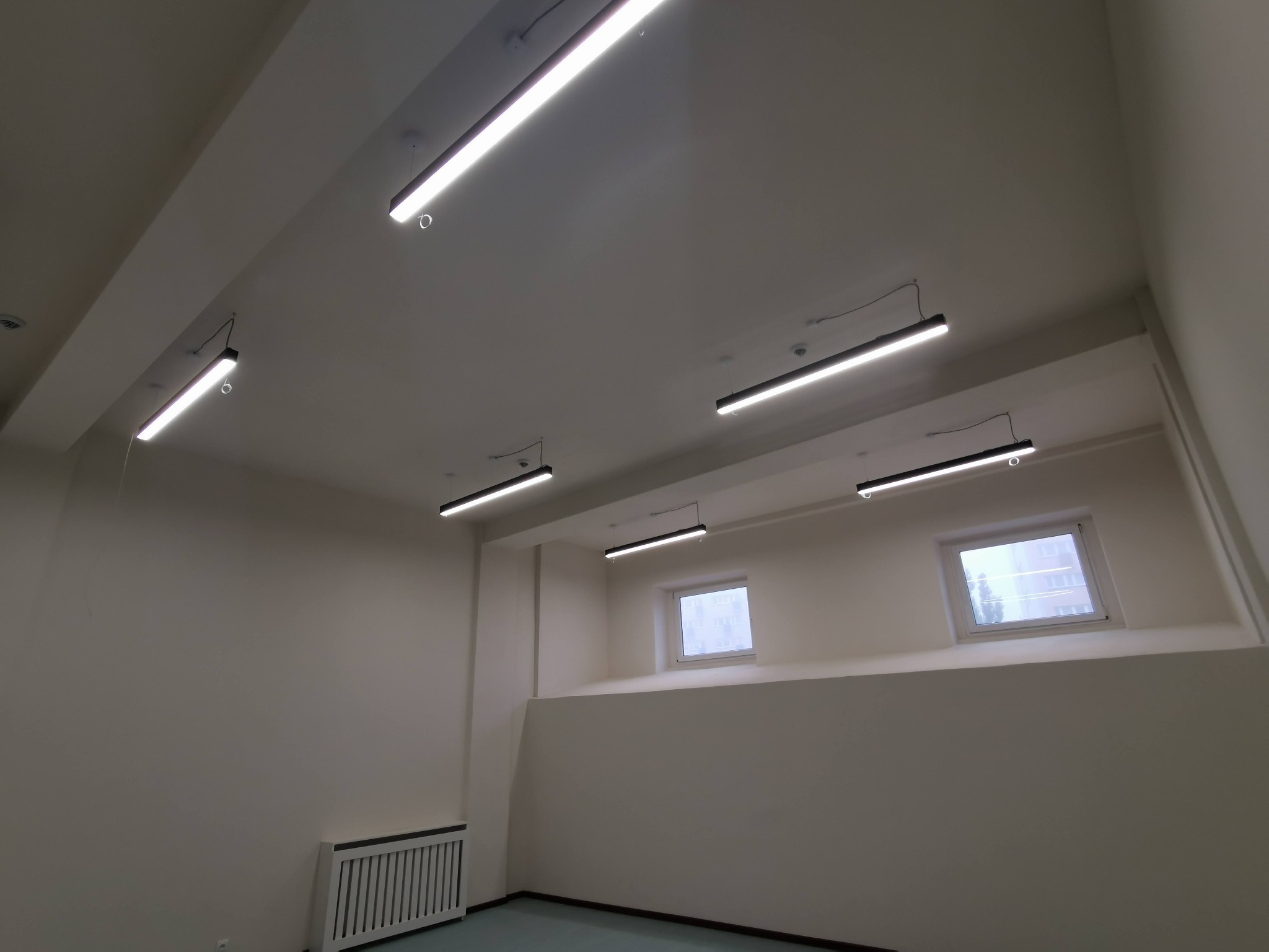 Wnętrze pomieszczenia z zamontowanymi liniowymi oprawami oświetleniowymi LED, widoczne okna z widokiem na budynek, białe ściany i grzejnik.