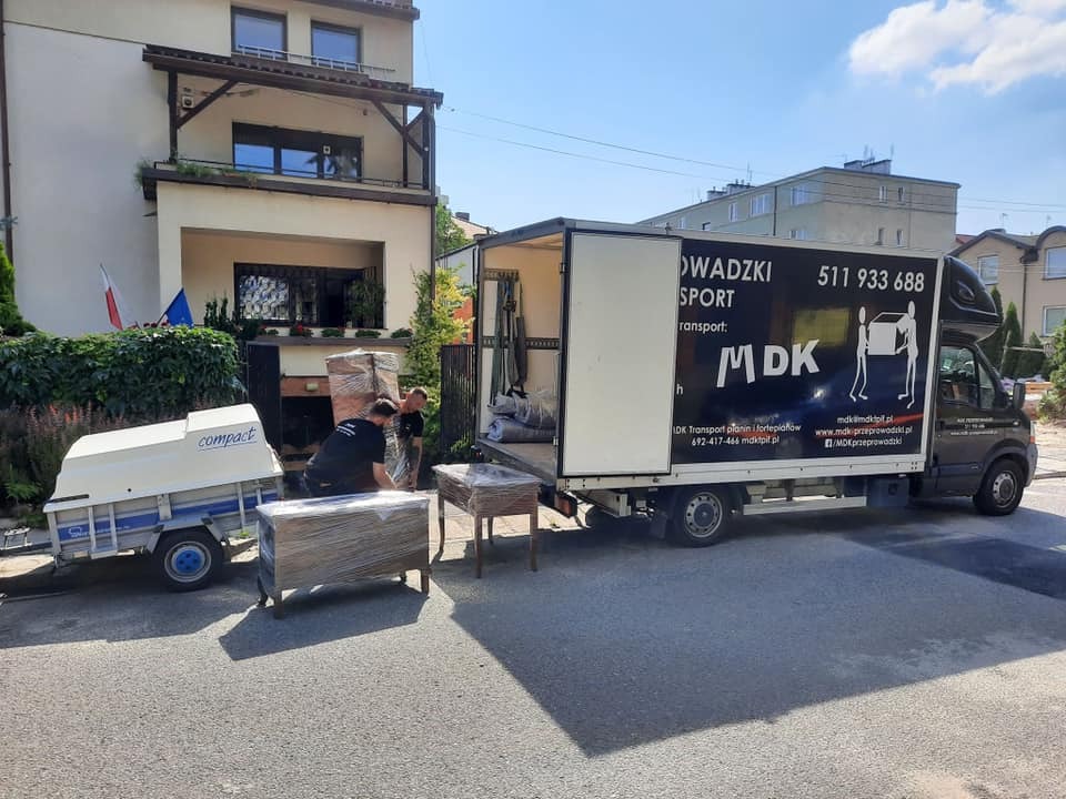 Ekipa przeprowadzkowa w Warszawie ładuje meble owinięte folią do samochodu dostawczego z logo firmy; widoczny również mały wózek transportowy.