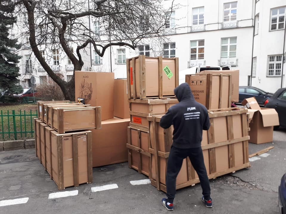 Mężczyzna w bluzie z kapturem stoi tyłem do obiektywu, obok stosu zabezpieczonych drewnianymi ramami kartonowych pudeł i kartonów na zewnątrz budynku.