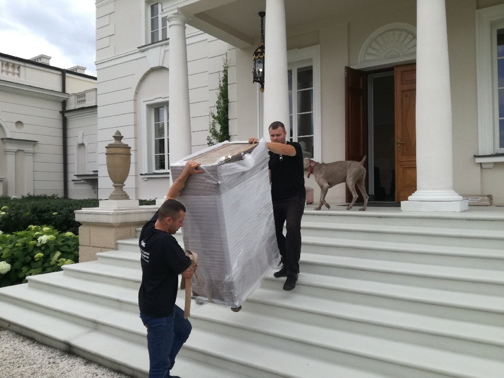 Dwóch mężczyzn w czarnych koszulkach wnosi mebel owinięty folią po marmurowych schodach do rezydencji, w tle pies rasy wyżeł.