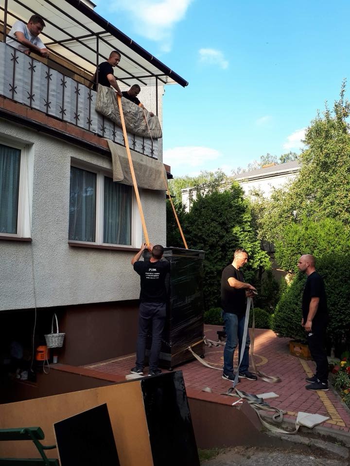 Przeprowadzka mebla z balkonu: czterech mężczyzn spuszcza pianino zabezpieczone folią i kocem za pomocą pasów transportowych z balkonu domu jednorodzinnego, dwóch na balkonie, dwóch na dole.