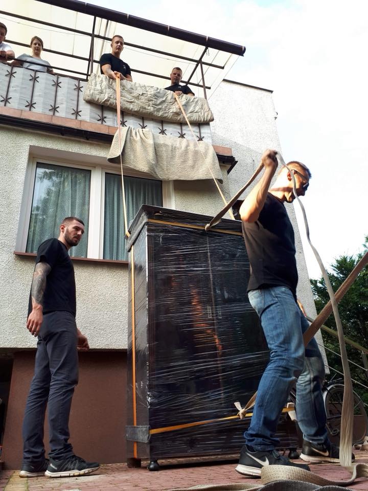 Ekipa spuszcza zabezpieczoną folią szafę z balkonu za pomocą pasów, jeden z pracowników nadzoruje proces na dole.