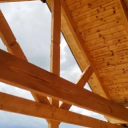 Drewniana konstrukcja dachu z widokiem na zielone wzg&oacute;rza i domy w oddali, jasne drewno, surowy stan, detale wykończeniowe na słupie.