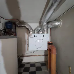 Biała centrala wentylacyjna z podłączonymi przewodami wentylacyjnymi w pomieszczeniu z czarno-białą podłogą w kratkę.