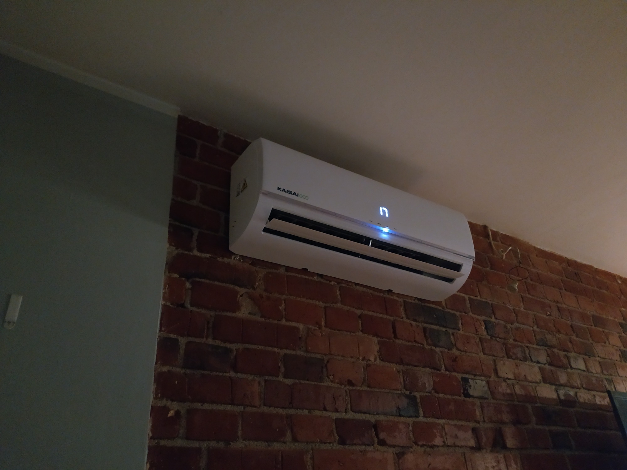 Biały klimatyzator Kaisai Eco zamontowany na ścianie z czerwonej cegły, wyświetlający temperaturę 17 stopni Celsjusza, widoczny fragment ściany pomalowanej na szaro.