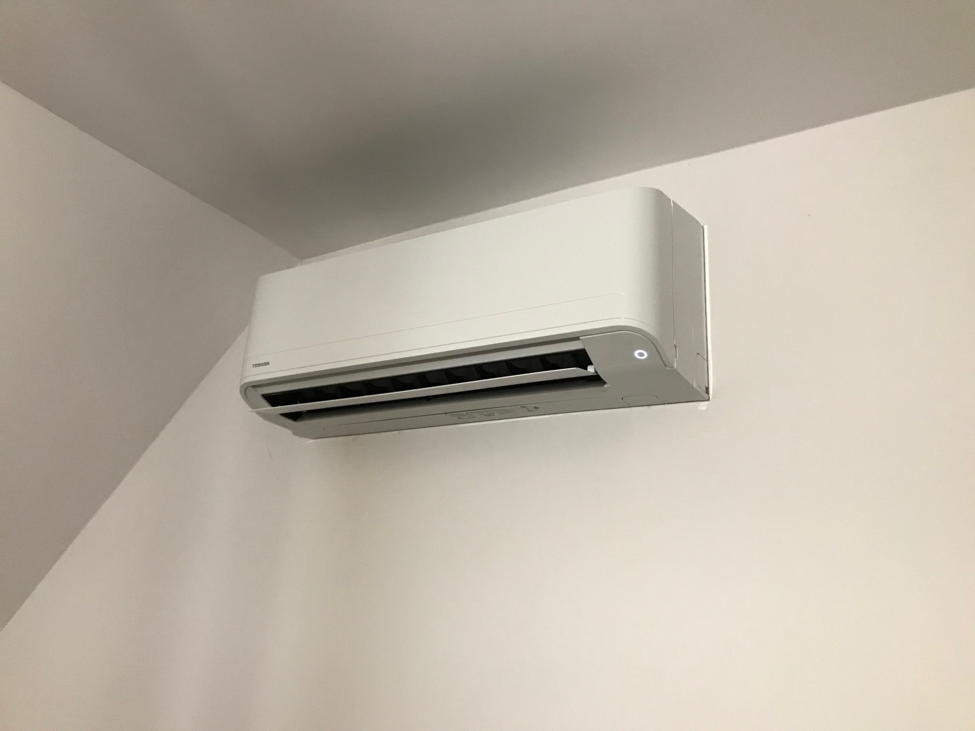 Biała jednostka klimatyzacji Toshiba zamontowana na białej ścianie, widoczna z boku, z delikatnym oświetleniem i cieniem na suficie w rogu pomieszczenia.
