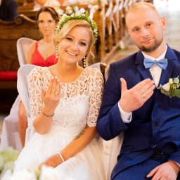 DreamArte Patrycja Stefaniak - Uśmiechnięta para młoda w kościele prezentuje obrączki, panna młoda w wianku, pan młody w niebieskim garniturze w kratę, w tle goście.