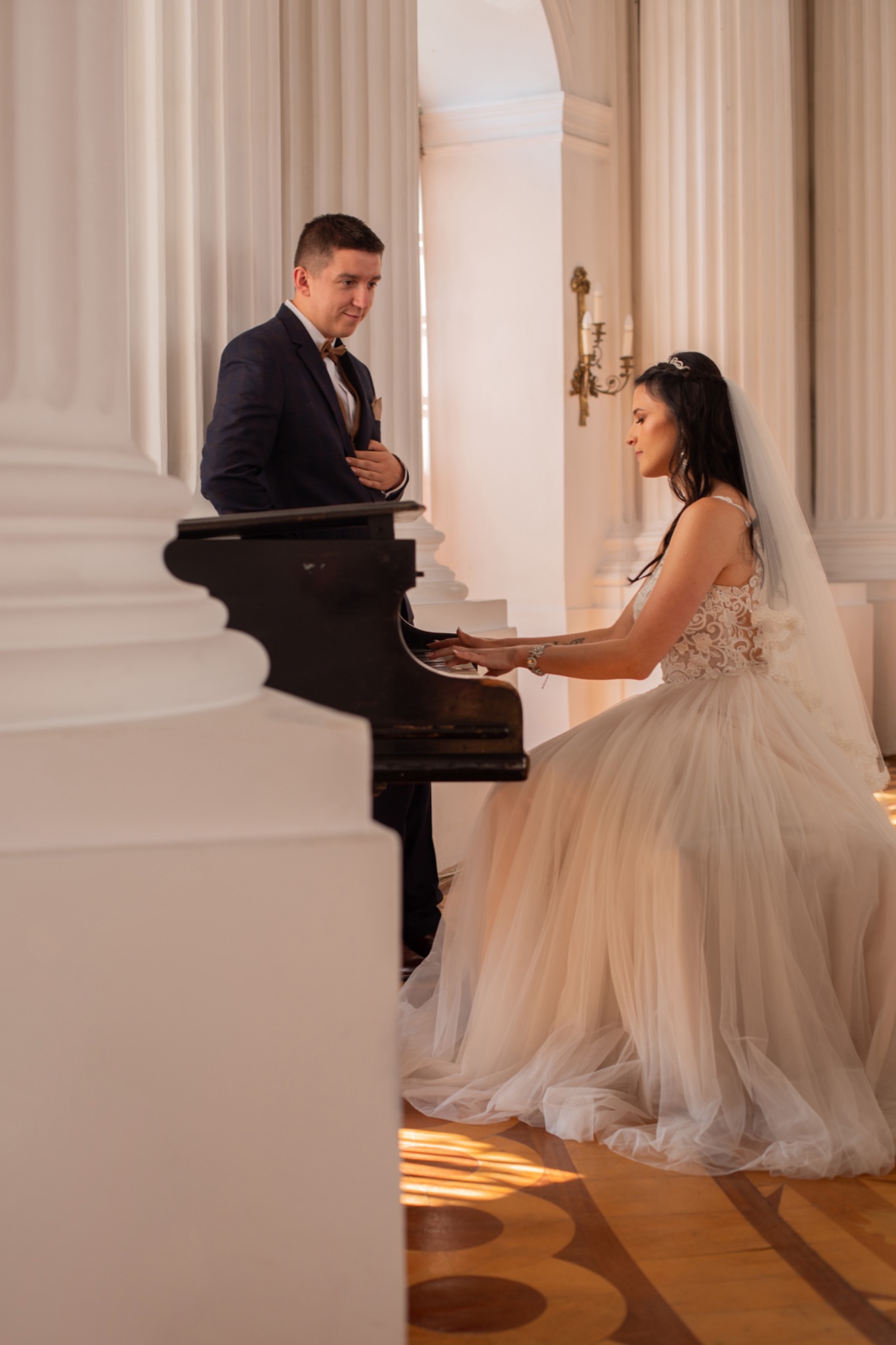 Panna młoda w sukni ślubnej gra na pianinie, obok stoi pan młody w ciemnym garniturze, jasne wnętrze z kolumnami.