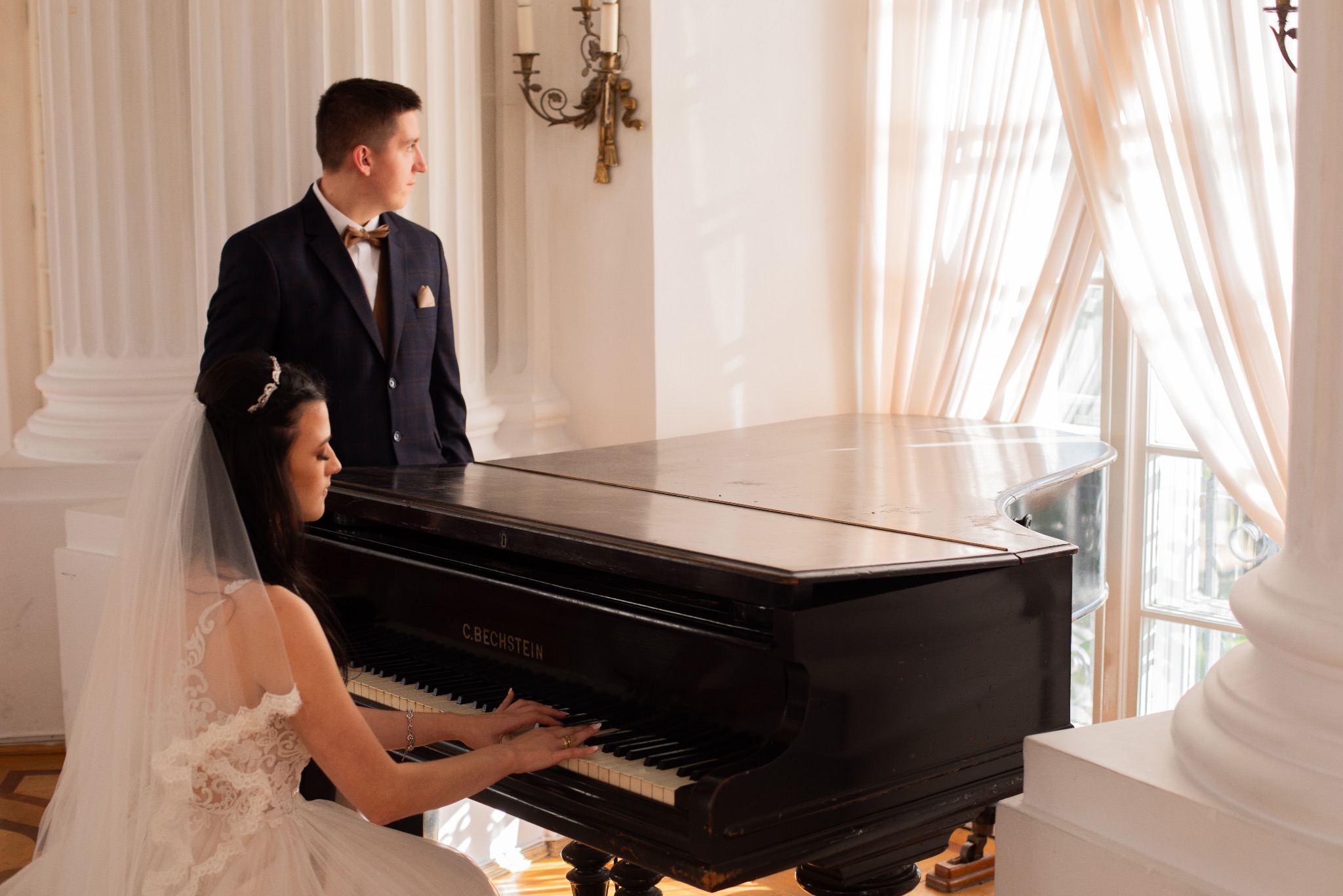 Panna młoda w białej sukni gra na czarnym fortepianie C. Bechstein, obok stoi pan młody w granatowym garniturze z muszką, jasne wnętrze z kolumnami i oknem w tle.