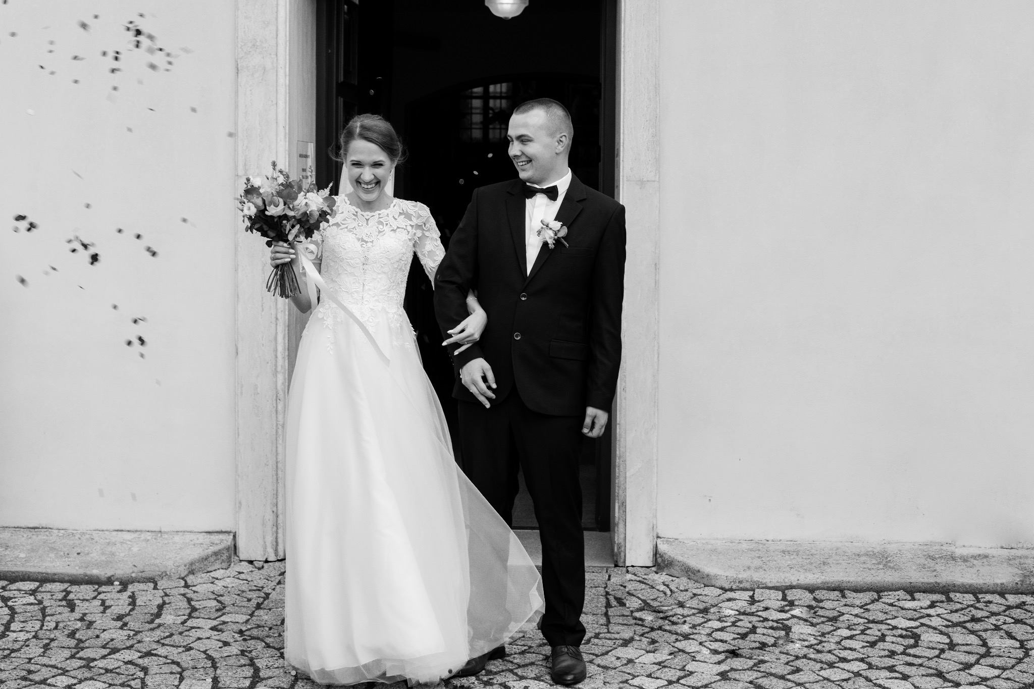 Czarno-białe zdjęcie uśmiechniętej pary młodej wychodzącej z kościoła, obsypywanej konfetti, panna młoda trzyma bukiet kwiatów.