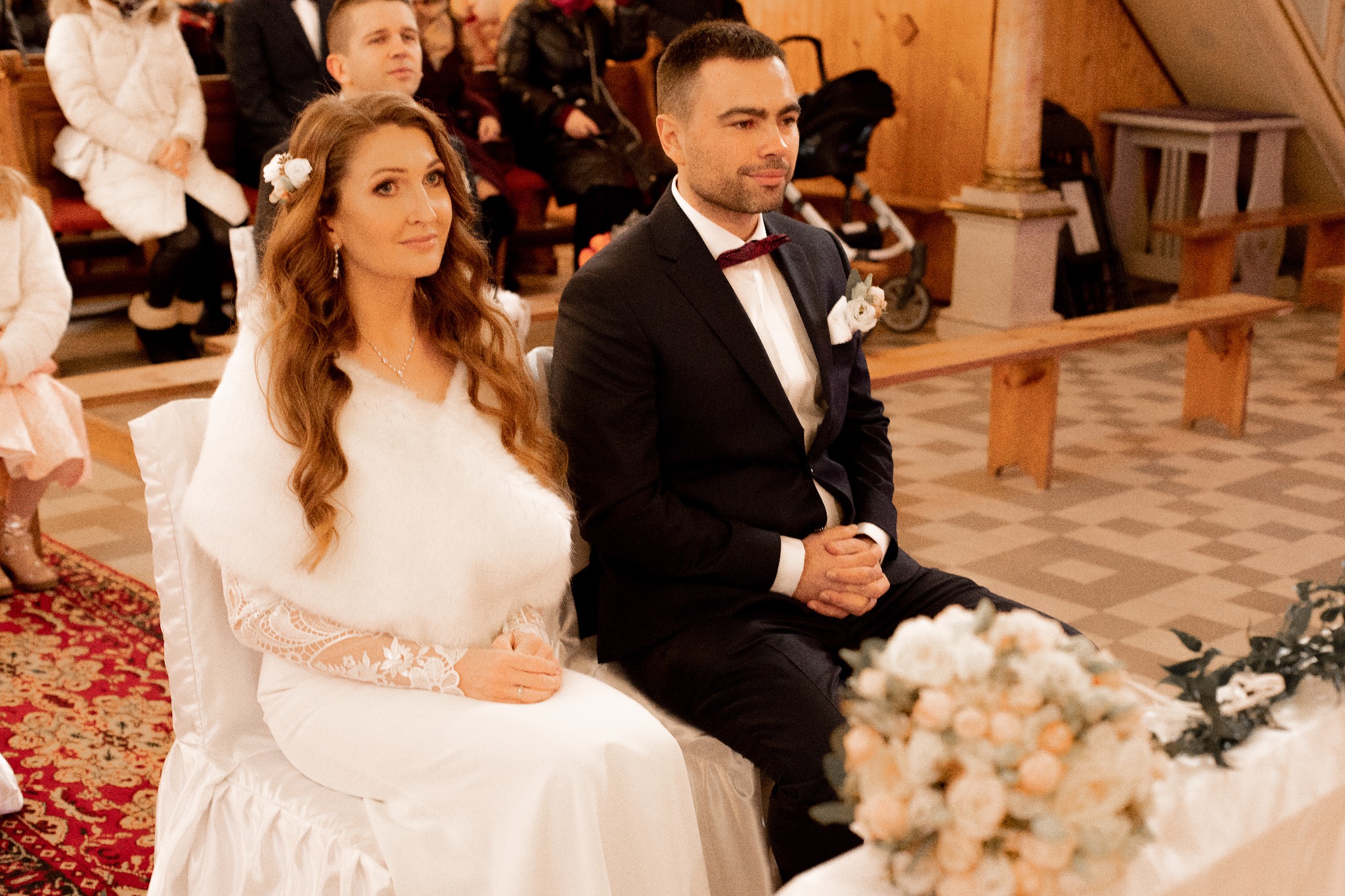 Para młoda siedzi w kościele podczas ceremonii ślubnej, panna młoda w białej sukni z futrzanym okryciem, pan młody w ciemnym garniturze z bordową muszką, w tle goście i dekoracje.