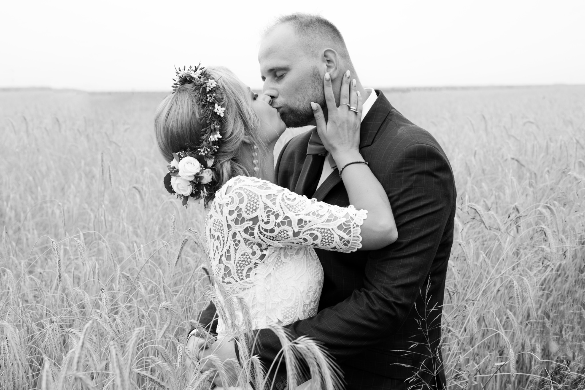 Czarno-białe zdjęcie całującej się pary młodej w polu zboża, panna młoda w koronkowej sukience i wianku, pan młody w ciemnym garniturze.