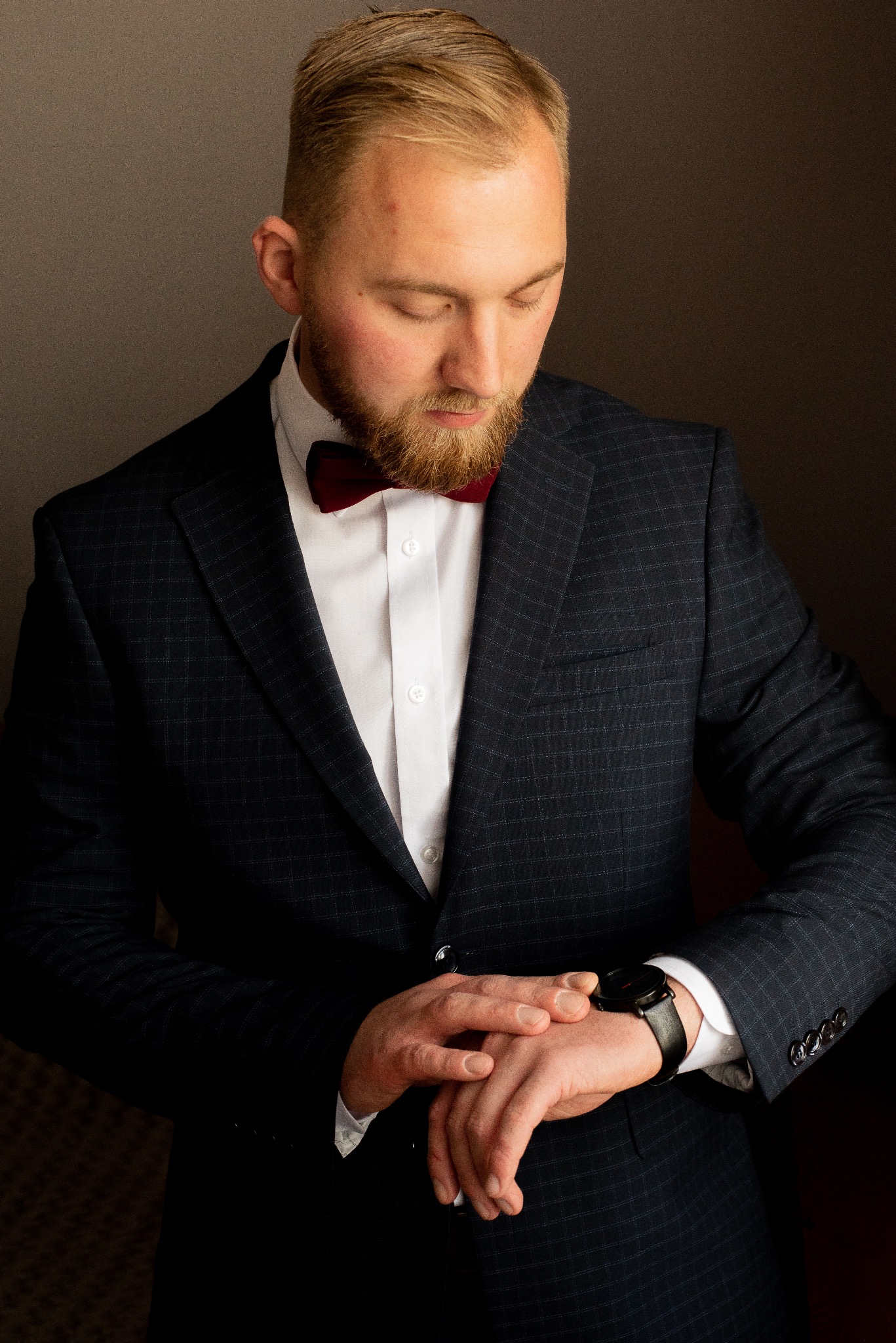 Mężczyzna w granatowym garniturze w kratę i bordowej muszce spogląda na zegarek na lewym nadgarstku, z bliska, w półmroku.