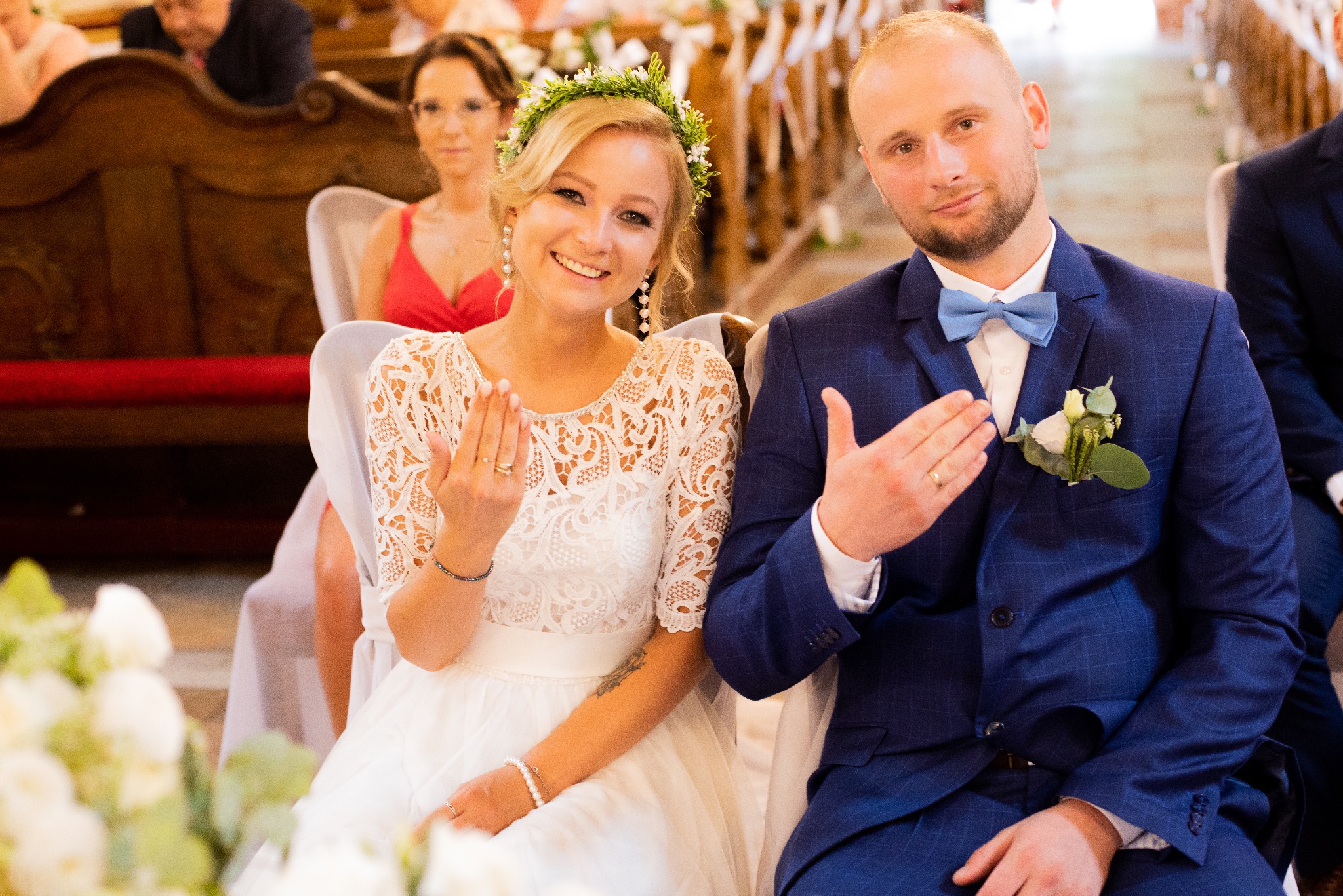 Uśmiechnięta para młoda w kościele prezentuje obrączki, panna młoda w wianku, pan młody w niebieskim garniturze w kratę, w tle goście.