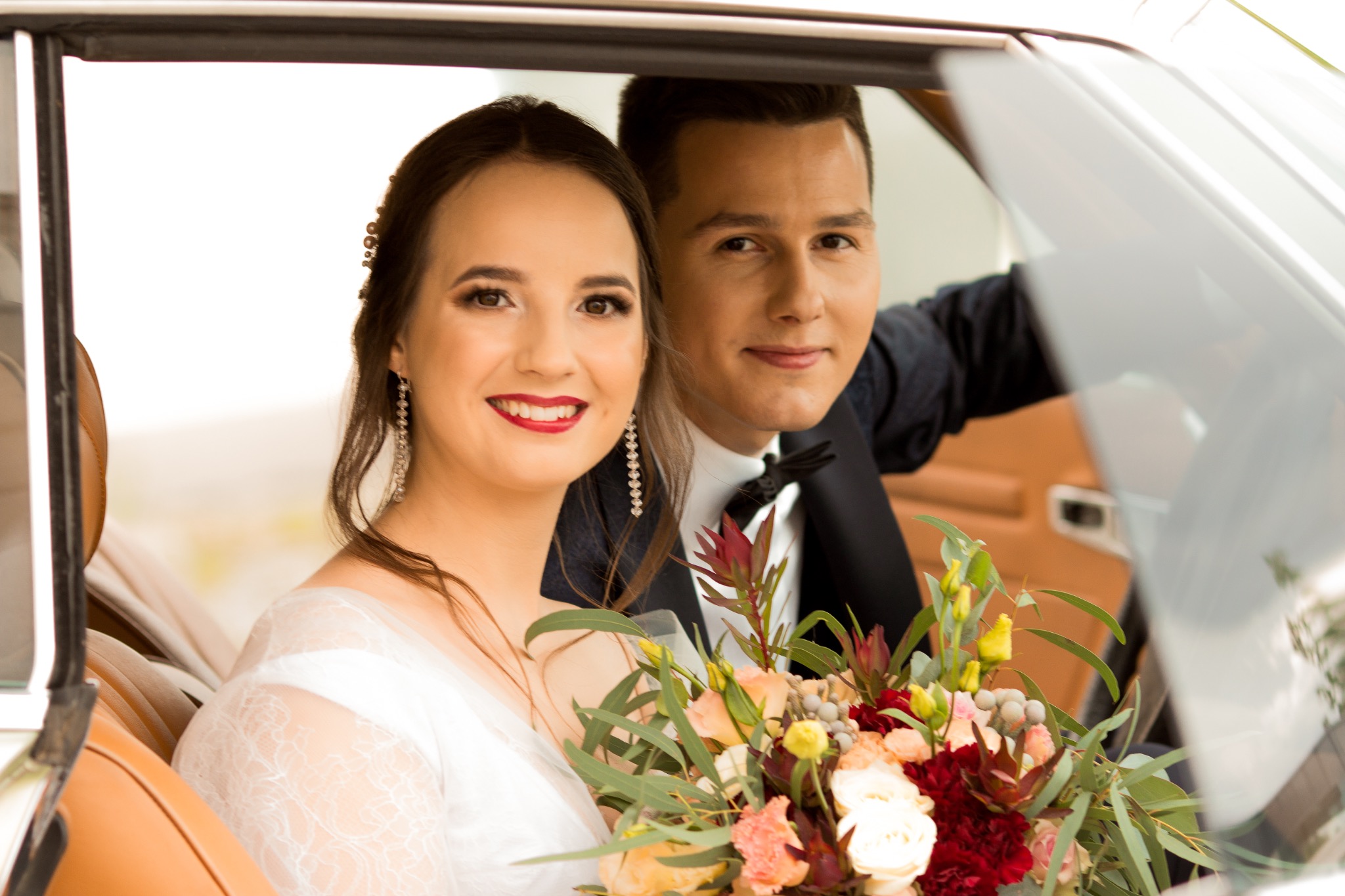 Uśmiechnięta para młoda w białym samochodzie z beżową skórzaną tapicerką, panna młoda trzyma bukiet z czerwonymi i pomarańczowymi kwiatami