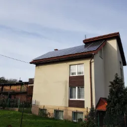 Dwukondygnacyjny dom z panelami słonecznymi na dachu pokrytym czerwoną dachówką, widok z poziomu gruntu, zieleń w ogrodzie i sąsiedni budynek w tle.