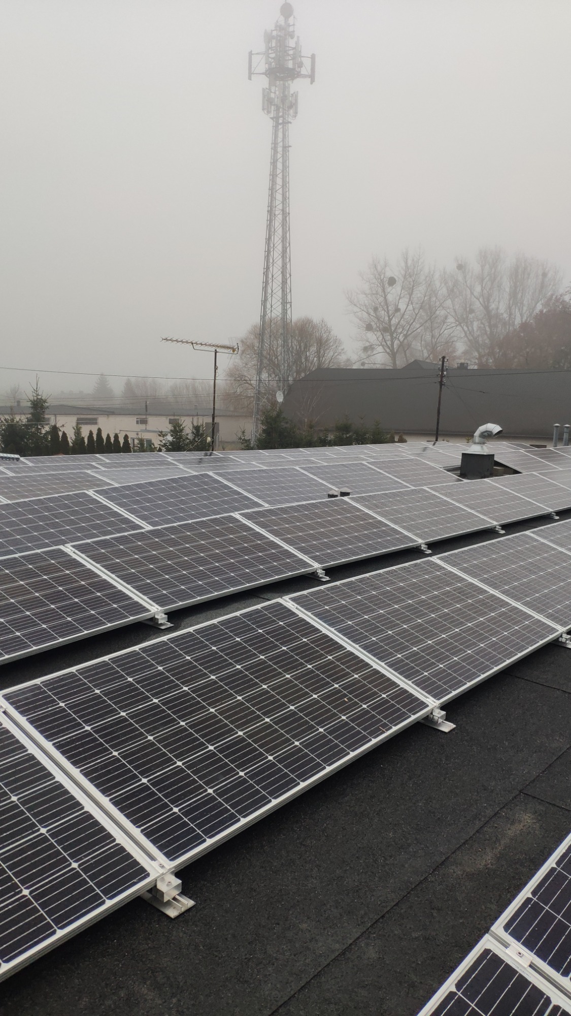 Rząd paneli słonecznych na dachu budynku w mglisty dzień, w tle maszt telefonii komórkowej.