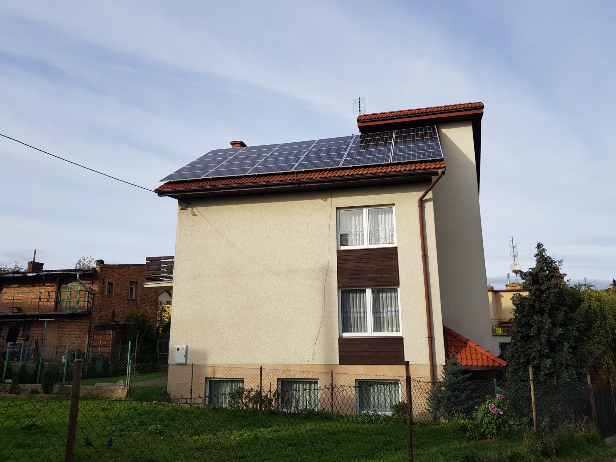 Dwukondygnacyjny dom z beżową elewacją i panelami słonecznymi na dachu z czerwonej dachówki, widok zza metalowego ogrodzenia na tle błękitnego nieba.