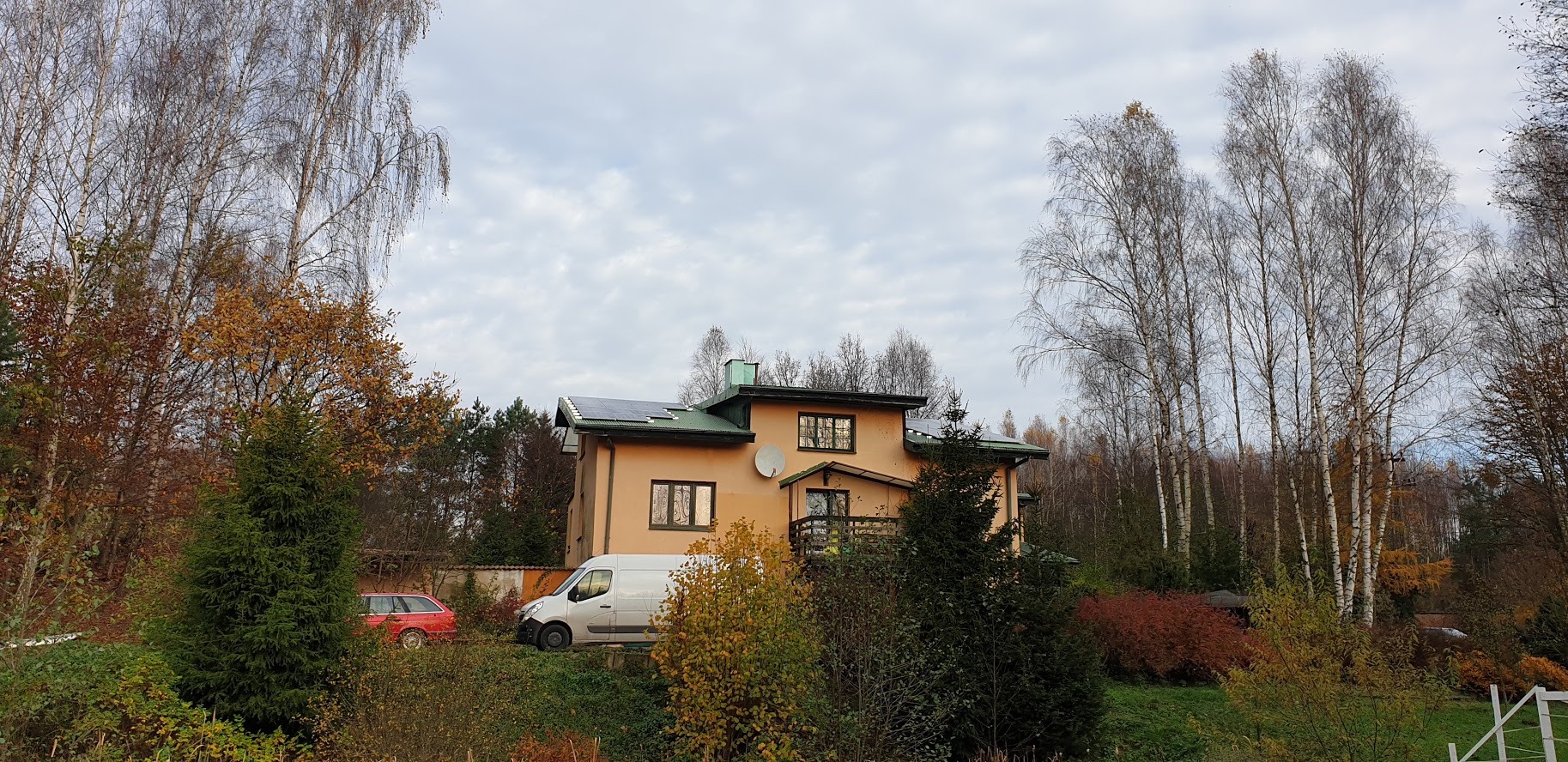 Dom jednorodzinny z zainstalowanymi panelami słonecznymi na dachu, widoczny biały samochód dostawczy i czerwone kombi na tle jesiennego krajobrazu.
