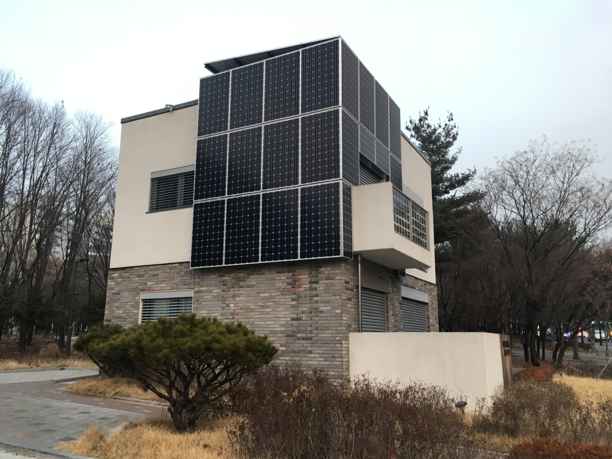 Dwukondygnacyjny budynek z ceglaną dolną częścią i jasną górną częścią, obłożony panelami słonecznymi na dachu i ścianach, otoczony drzewami i krzewami w jesiennej scenerii.