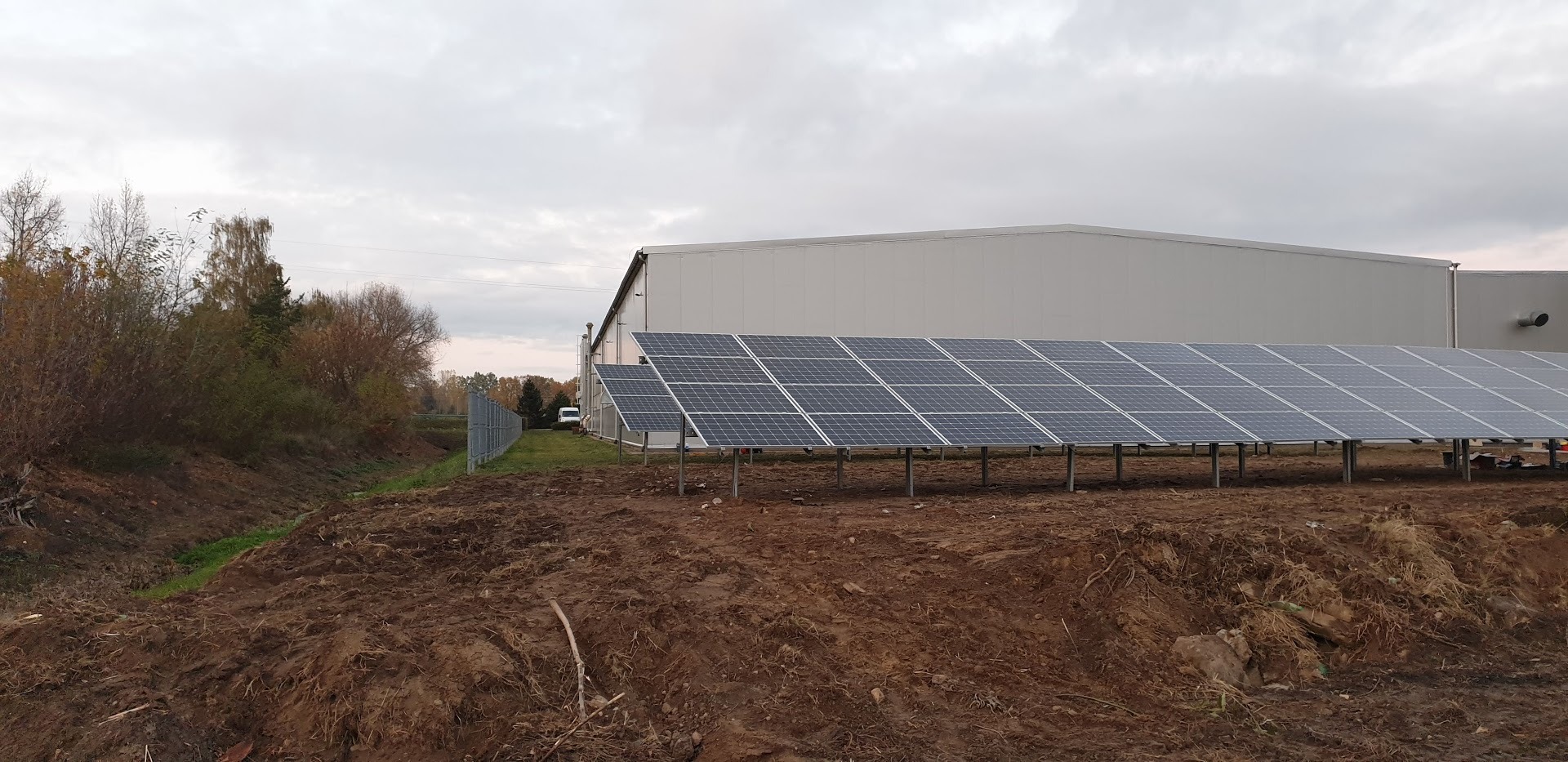 Rząd paneli słonecznych na metalowej konstrukcji, zainstalowanych na ziemi obok budynku magazynowego w pochmurny dzień.