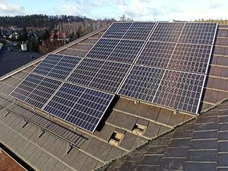 Panele słoneczne zainstalowane na brązowym dachu z widocznymi ubytkami w pokryciu, w tle zalesione wzgórze.