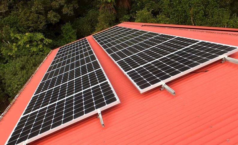 Dwa panele fotowoltaiczne zamontowane na czerwonym, trapezowym dachu z widocznymi mocowaniami i zielenią drzew w tle.
