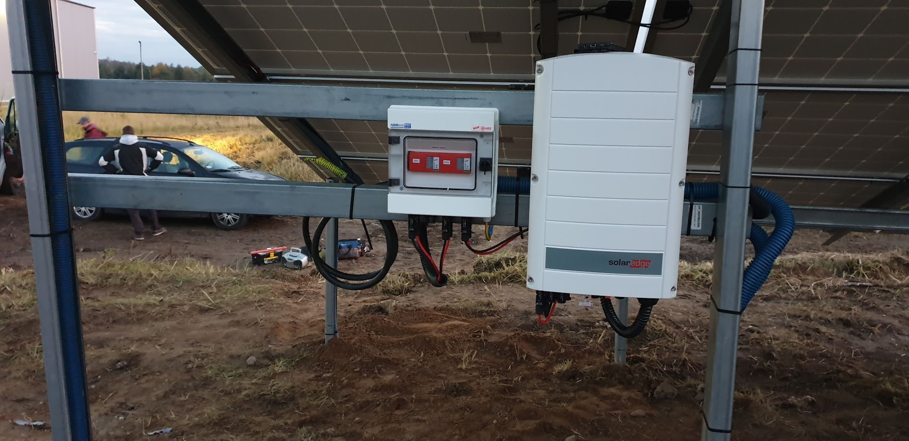 Instalacja paneli słonecznych w trakcie montażu: widoczny inwerter SolarEdge, skrzynka z zabezpieczeniami, okablowanie oraz konstrukcja wsporcza na tle ziemi i samochodu.