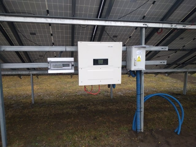 Widok od spodu na panele słoneczne z widocznym inwerterem, skrzynką elektryczną i niebieskimi rurami ochronnymi na tle trawnika.