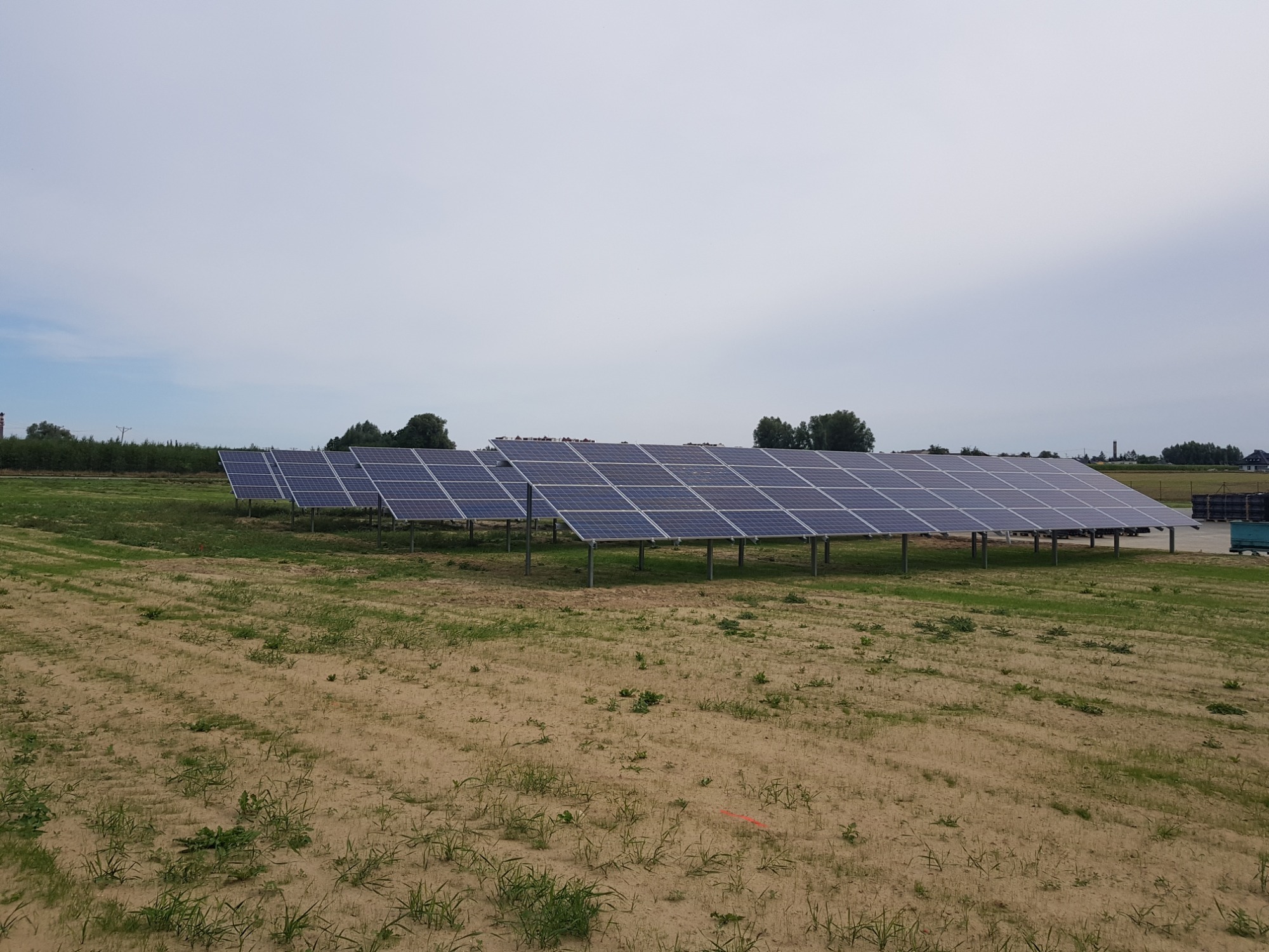 Rząd paneli fotowoltaicznych na metalowych konstrukcjach, ustawionych na piaszczystym polu z rzadką roślinnością, pod pochmurnym niebem.