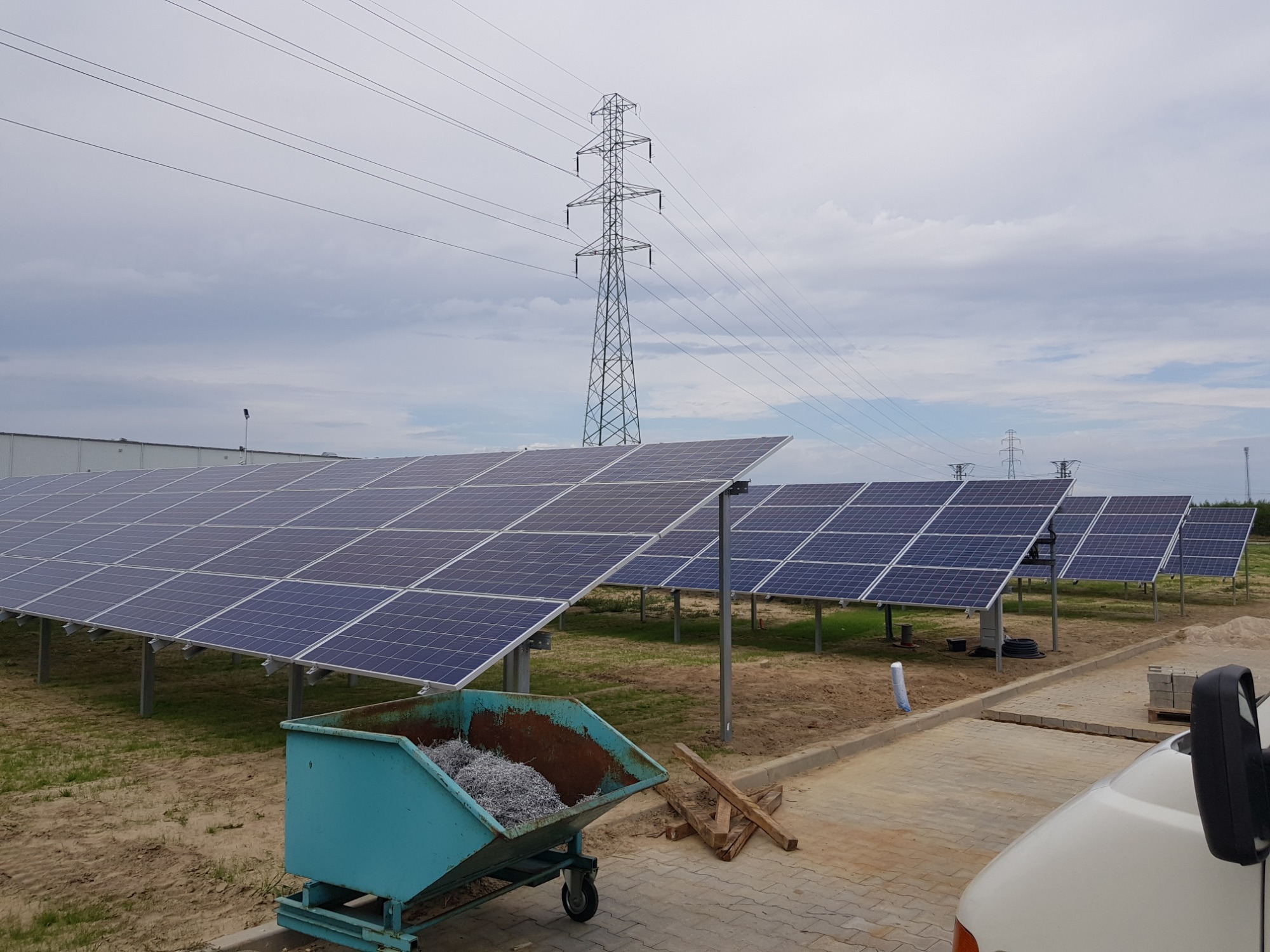 Rzędy paneli słonecznych na metalowych konstrukcjach, niebieski kontener wypełniony odpadami, słup wysokiego napięcia w tle, widok zza samochodu.