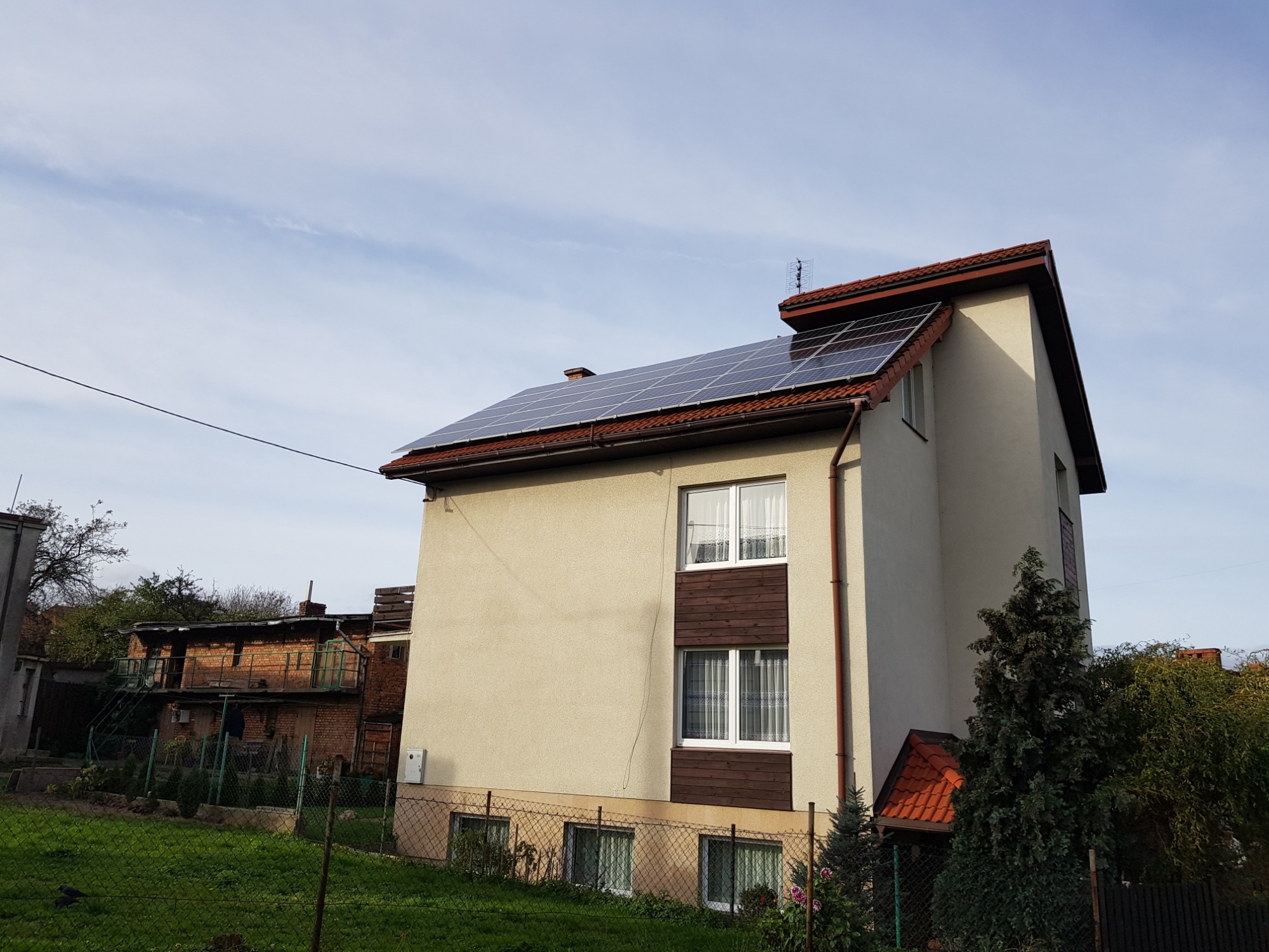 Dwukondygnacyjny dom z panelami słonecznymi na dachu pokrytym czerwoną dachówką, widok z poziomu gruntu, zieleń w ogrodzie i sąsiedni budynek w tle.