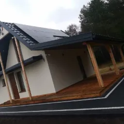 Nowoczesny dom z panelami słonecznymi, drewnianym tarasem i czarną nawierzchnią z białymi obramowaniami, widoczny ubijak wibracyjny.