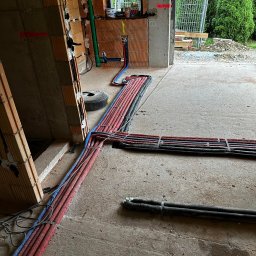 Kopitza Instalacje - Wnętrze budynku w trakcie instalacji hydraulicznej i grzewczej: liczne rury w kolorach czerwonym, niebieskim i czarnym prowadzone po posadzce, ściany z pustaków ceramicznych z widocznymi...