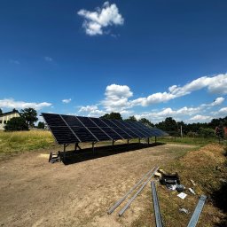 Kopitza Instalacje - Rozległa instalacja fotowoltaiczna na metalowej konstrukcji w trakcie montażu, widoczne narzędzia i elementy montażowe na ziemi, błękitne niebo z chmurami w tle.