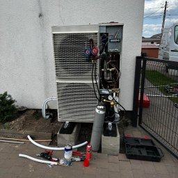 Kopitza Instalacje - Montaż zewnętrznej jednostki pompy ciepła z otwartą obudową, widoczne elementy instalacji, butla z gazem chłodniczym, narzędzia na kostce brukowej, budynek w tle.