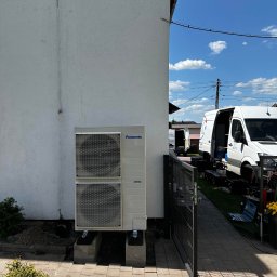 Kopitza Instalacje - Zewnętrzna jednostka pompy ciepła Panasonic zamontowana przy białej ścianie domu, widoczne dwa samochody serwisowe firmy w tle, brama wejściowa i narzędzia.