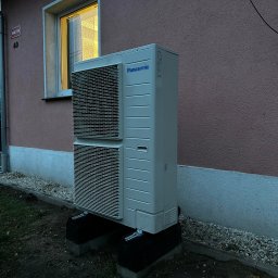 Kopitza Instalacje - Zewnętrzna jednostka pompy ciepła Panasonic zamontowana na betonowych podstawach przy ścianie budynku z oknem, widoczne elementy instalacji.