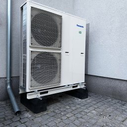 Kopitza Instalacje - Zewnętrzna jednostka pompy ciepła Panasonic R32, zamontowana na czarnych podstawach z szarej kostki brukowej, widoczny fragment rury spustowej przy ścianie budynku z szarym tynkiem.