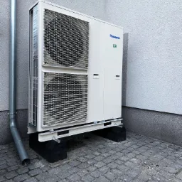 Zewnętrzna jednostka pompy ciepła Panasonic R32, zamontowana na czarnych podstawach z szarej kostki brukowej, widoczny fragment rury spustowej przy ścianie budynku z szarym tynkiem.