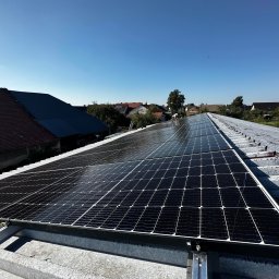 Kopitza Instalacje - Panele fotowoltaiczne zamontowane na blaszanym dachu w wiejskiej scenerii, widoczne domy z czerwonymi dachówkami i pracownik w oddali.