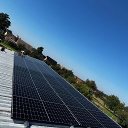 Kopitza Instalacje - Instalacja paneli słonecznych na blaszanym dachu budynku w słoneczny dzień, widok z dołu pod kątem, pracownik w oddali przy panelach.