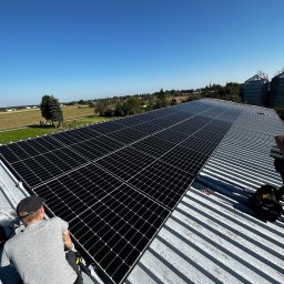 Kopitza Instalacje - Monter w szarej koszulce instaluje panele słoneczne na blaszanym dachu; w tle wieś i silosy zbożowe pod błękitnym niebem.