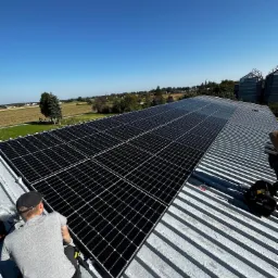 Monter w szarej koszulce instaluje panele słoneczne na blaszanym dachu; w tle wieś i silosy zbożowe pod błękitnym niebem.