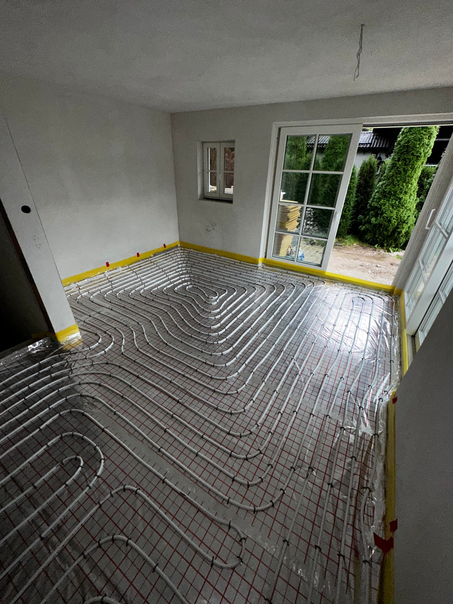 Białe rurki ogrzewania podłogowego ułożone w pętle na folii z czerwoną siatką, przyklejone żółtą taśmą do ścian w jasnym pomieszczeniu z oknem balkonowym wychodzącym na zieleń.